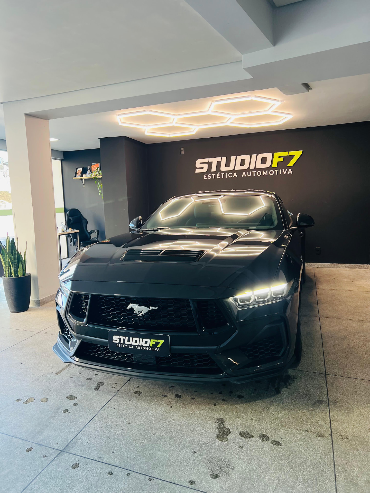 Ford Mustang preto no box da StudioF7 sob luminária hexagonal, com logo ao fundo
